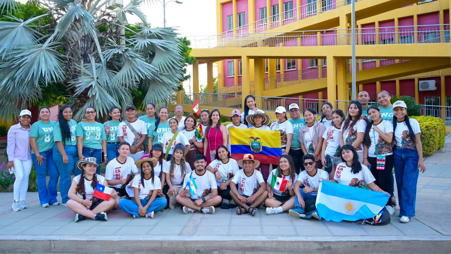Grupo de aproximadamente 30 jóvenes posando frente a un edificio amarillo y rosa, sosteniendo banderas de varios países latinoamericanos (Ecuador, México, Argentina y Chile), muchos vistiendo camisetas blancas con logos, con palmeras al fondo.