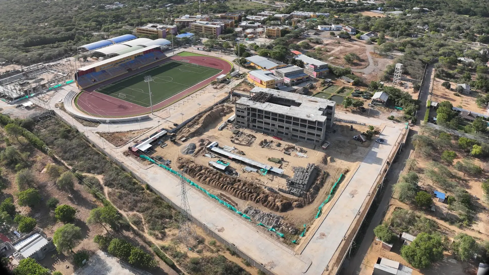 Vista aérea del campus universitario que muestra una obra en construcción de un edificio multi-piso, junto al estadio con cancha de fútbol y pista de atletismo, rodeados de áreas verdes y vegetación local.