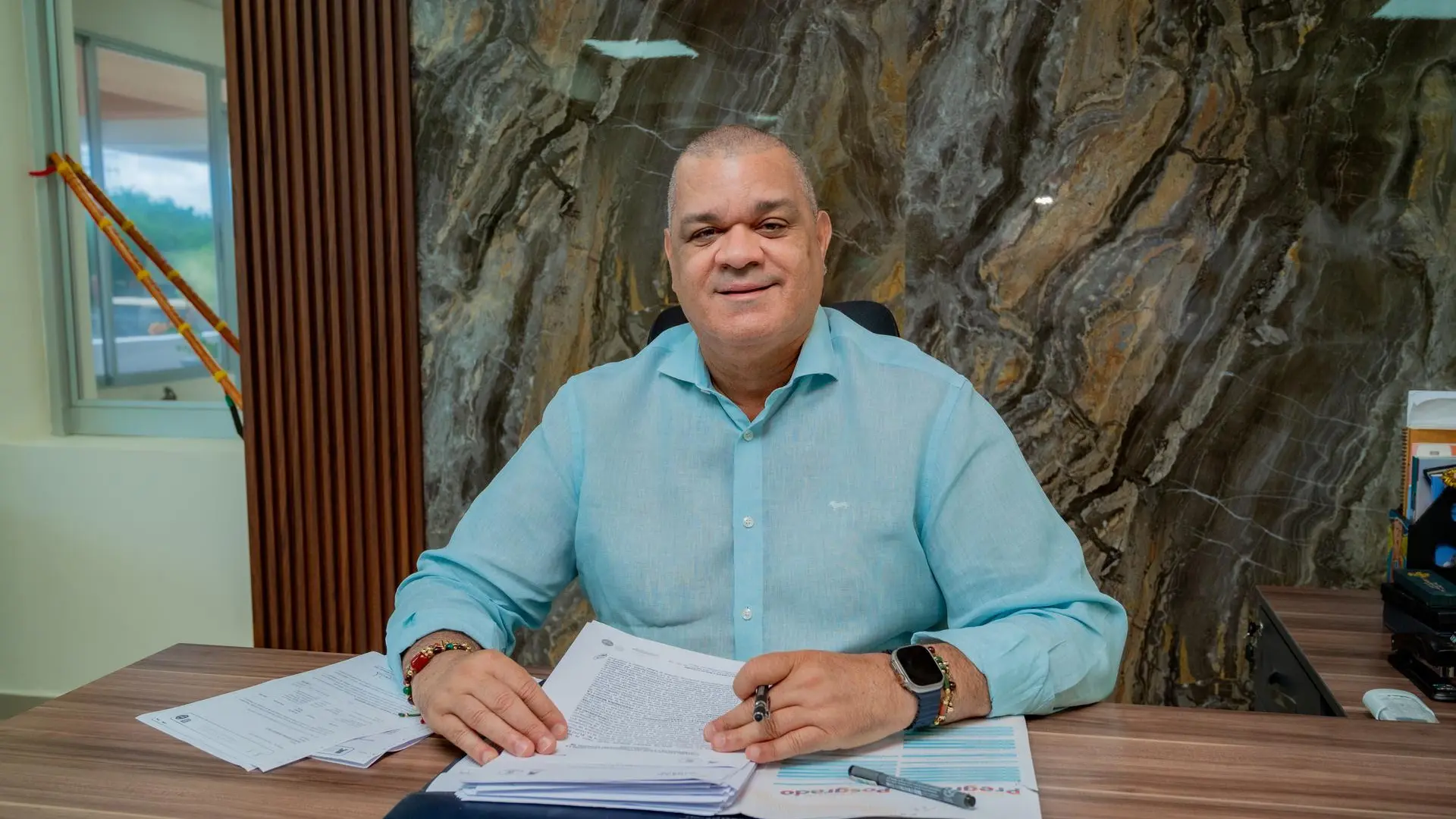 Hombre sentado detrás de un escritorio, sonríe a la cámara mientras sostiene documentos y un bolígrafo. Viste una camisa azul claro y está en una oficina con un fondo de mármol oscuro y elementos decorativos.