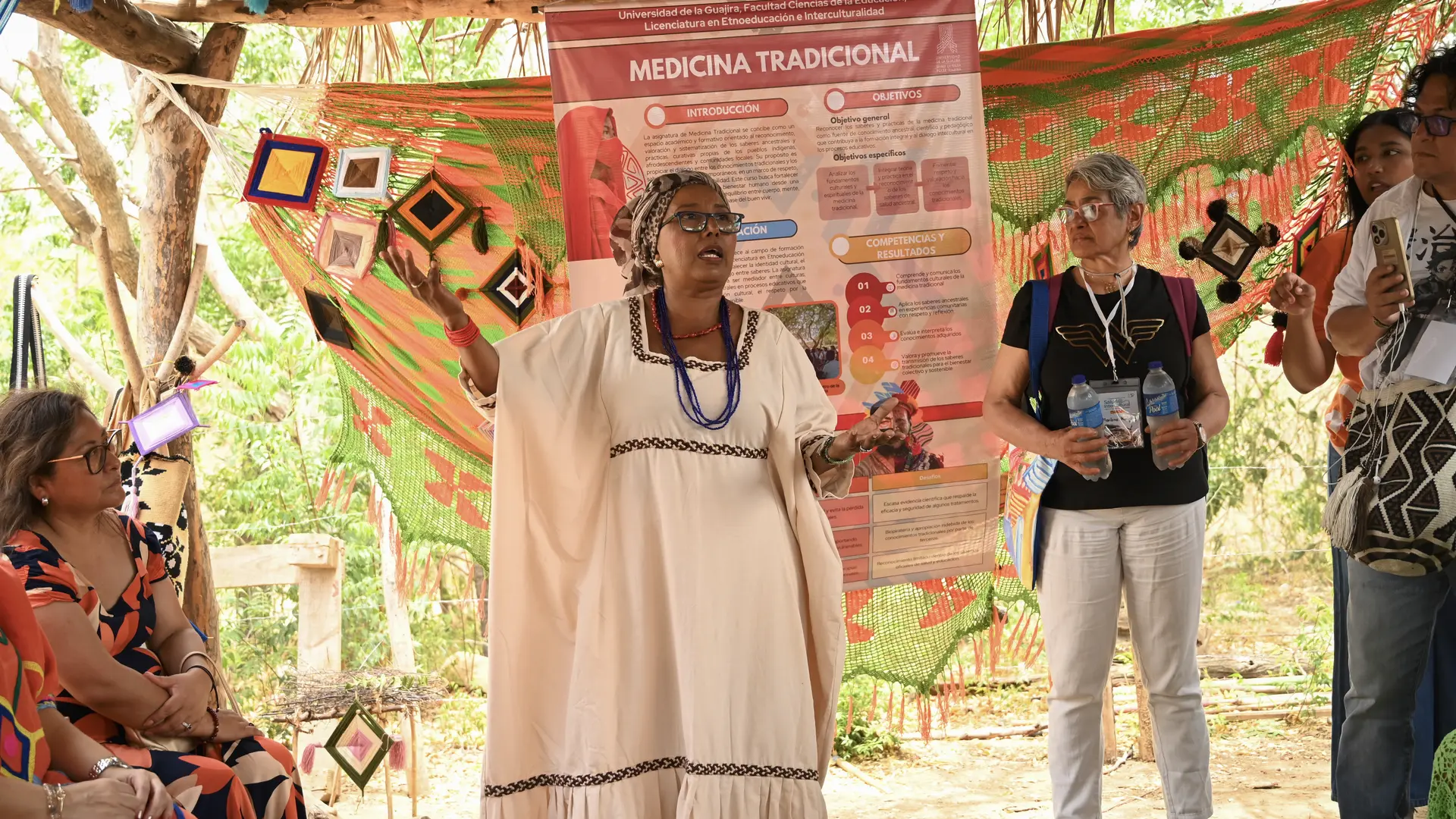 En un espacio al aire libre bajo una enramada tradicional con chinchorros de colores de fondo, una mujer indígena wayuu ataviada con manta guajira beige, turbante y collares oscuros habla y expone gesticulando con las manos abiertas. Detrás de ella se observa un póster informativo de la Universidad de la Guajira titulado "Medicina Tradicional".
