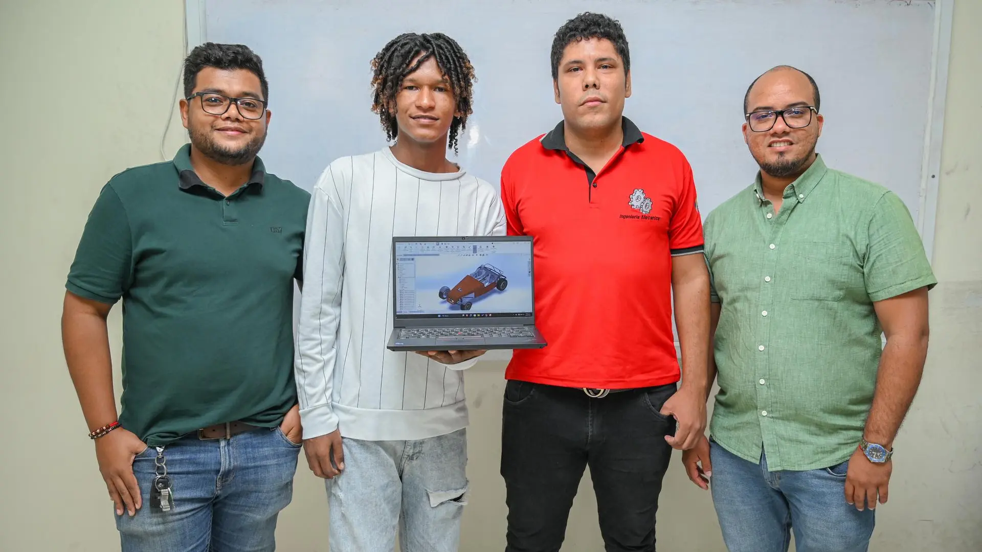 Cuatro personas posan frente a un tablero en un aula,mientras uno de ellos sostiene un computador portátil que muestra el diseño 3D de un vehiculo