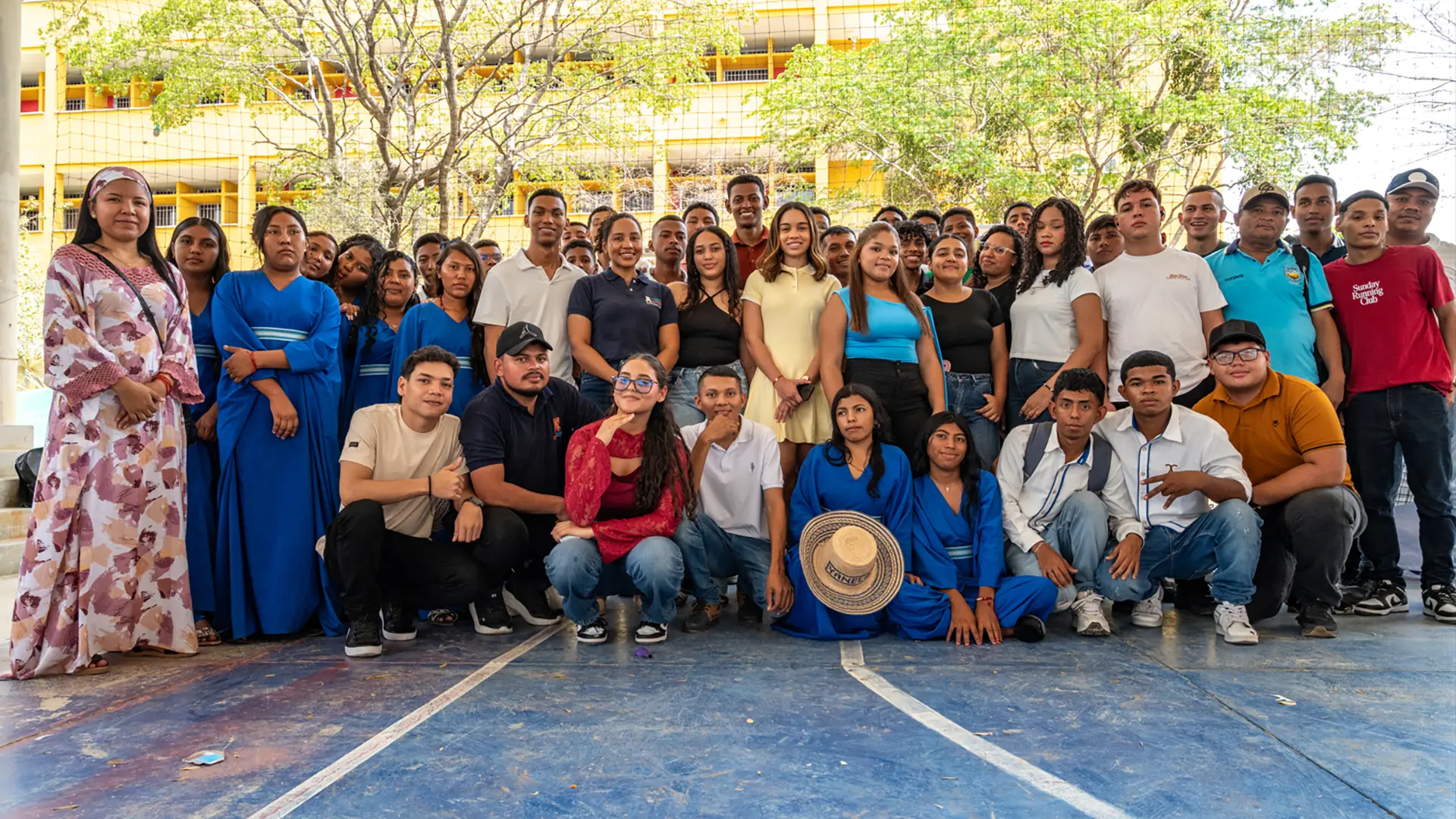 Grupo de estudiantes reunidos y posando para una fotografía en una cancha deportiva, frente a un edificio universitario. Varias personas visten atuendos tradicionales y prendas de diferentes colores.