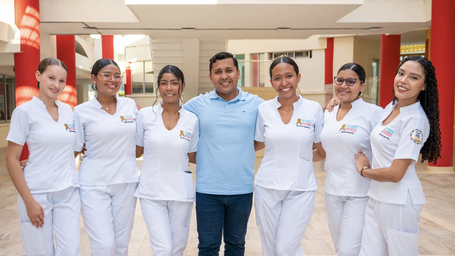 Grupo de seis estudiantes de enfermería vestidas con uniforme blanco posa alegremente junto a su docente en un patio institucional.