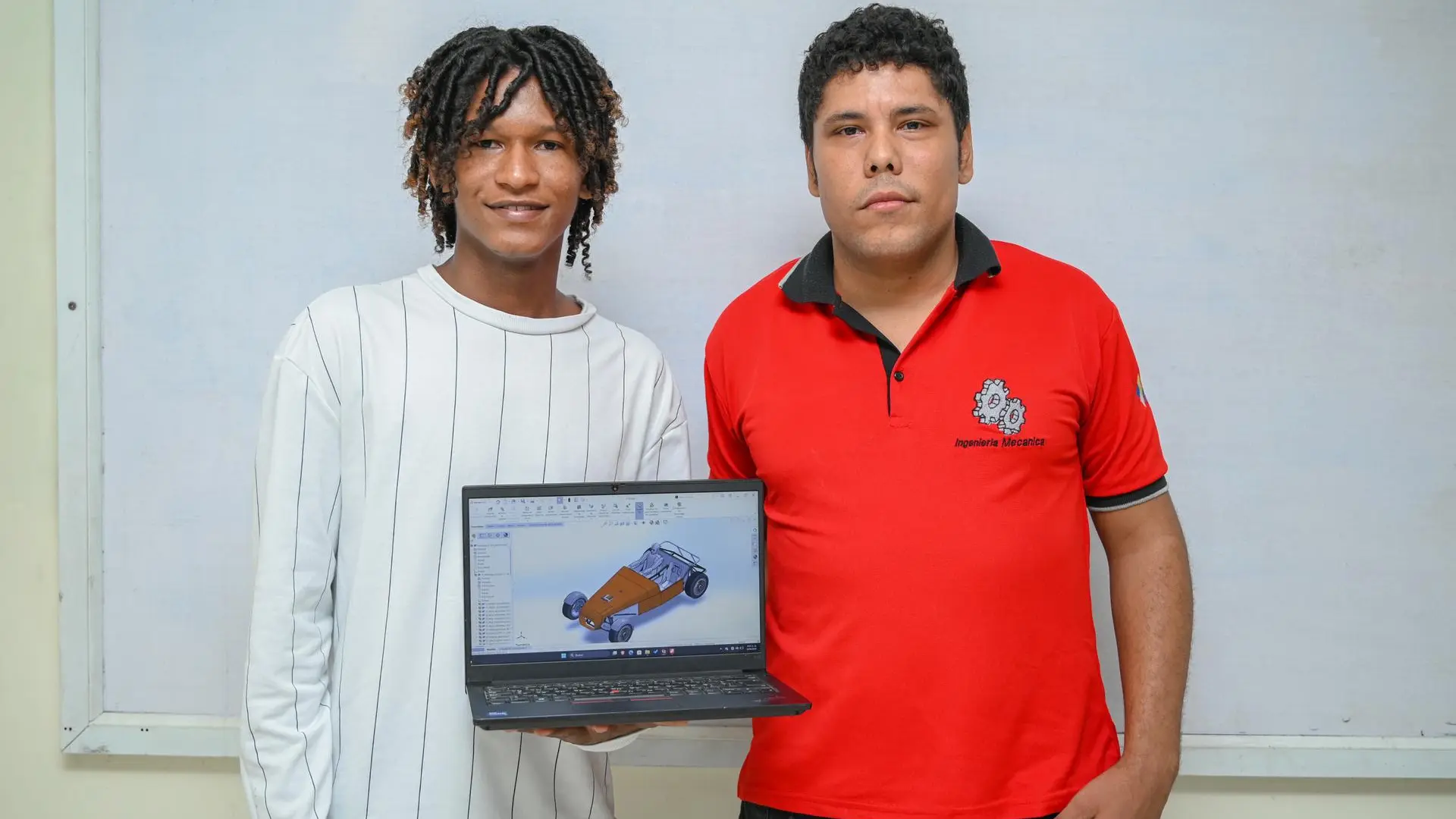 Dos personas posan frente a un tablero blanco mientras sostienen un computador portátil que muestra un diseño 3D de un vehículo realizado en software de modelado. La persona de la derecha viste una camiseta roja con el logo de Ingeniería Mecánica, resaltando una actividad académica relacionada con diseño asistido por computadora y formación en ingeniería.