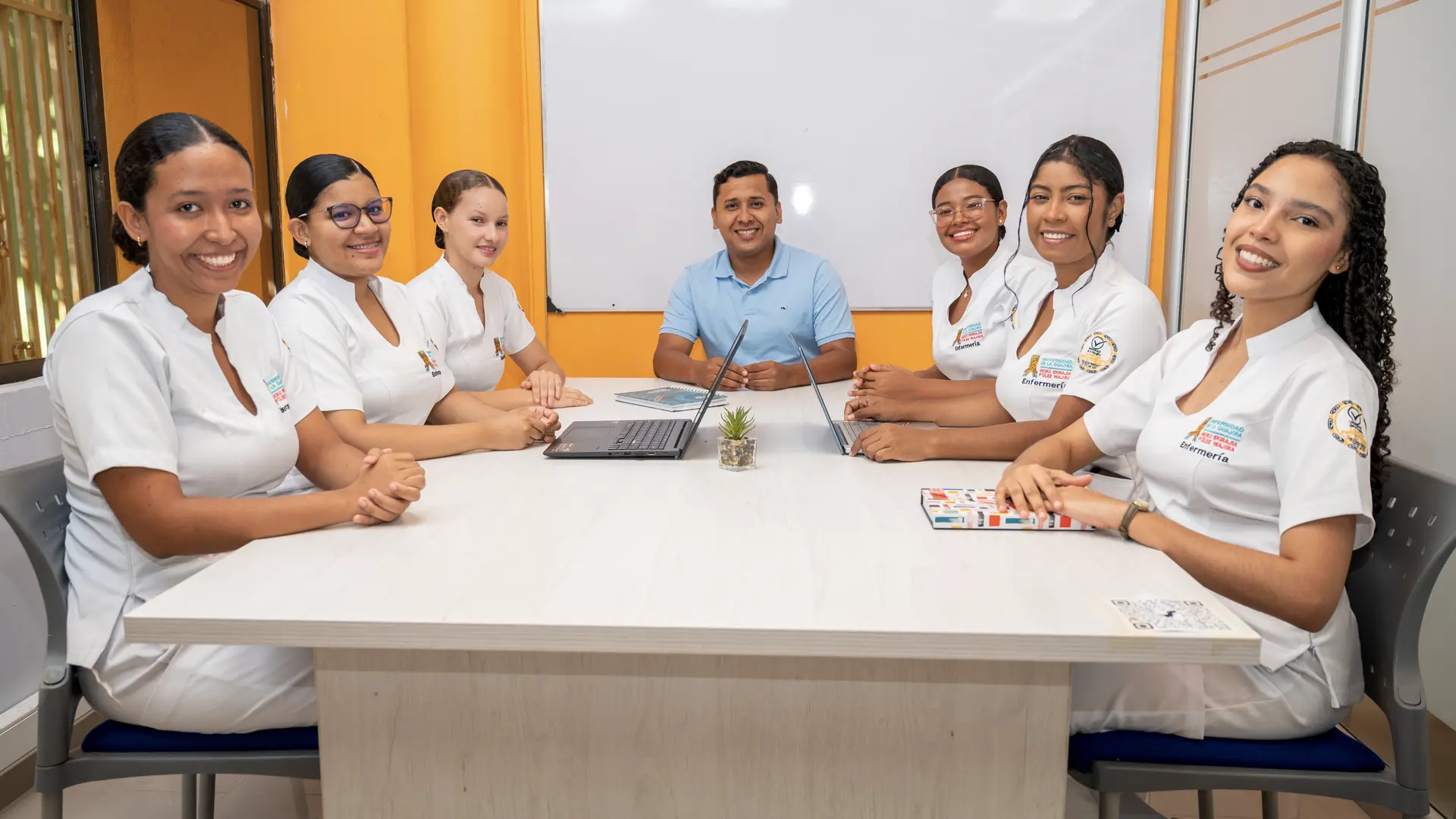 Seis estudiantes de enfermería con uniformes blancos y un docente con camiseta azul claro están sentados alrededor de una mesa de reuniones rectangular en un aula o sala de estudio con paredes naranja y una pizarra blanca al fondo. Las estudiantes, distribuidas a ambos lados, y el profesor, en el centro, sonríen a la cámara. En la mesa se observan un par de laptops, libretas y una pequeña planta.