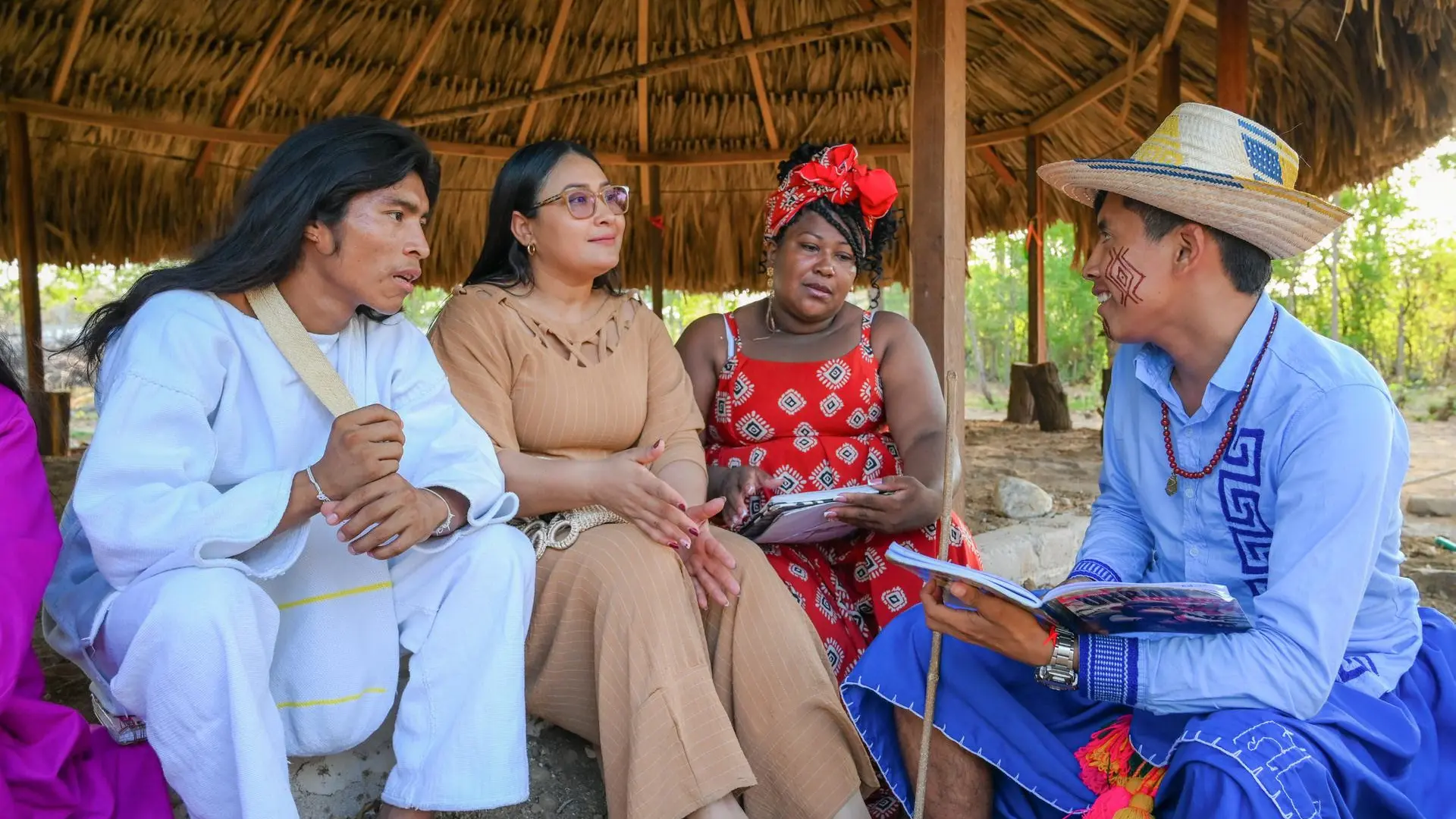Grupo de cuatro personas sentadas bajo un techo de paja durante lo que parece ser una reunión comunitaria o sesión de capacitación. Las personas visten ropa tradicional e indígena con diseños y colores distintivos, algunos con accesorios y adornos tradicionales. Sostienen documentos y materiales educativos.