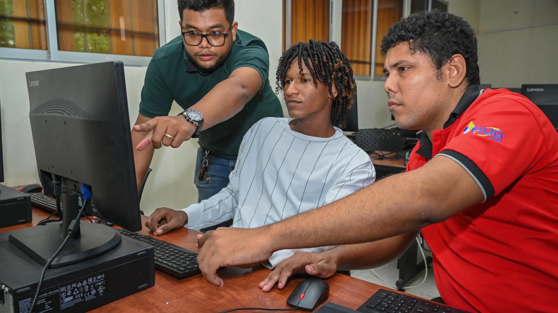 Tres personas trabajan juntas frente a un computador en un laboratorio de informática,revisando información en la pantalla y colaborando en una actividad académica.