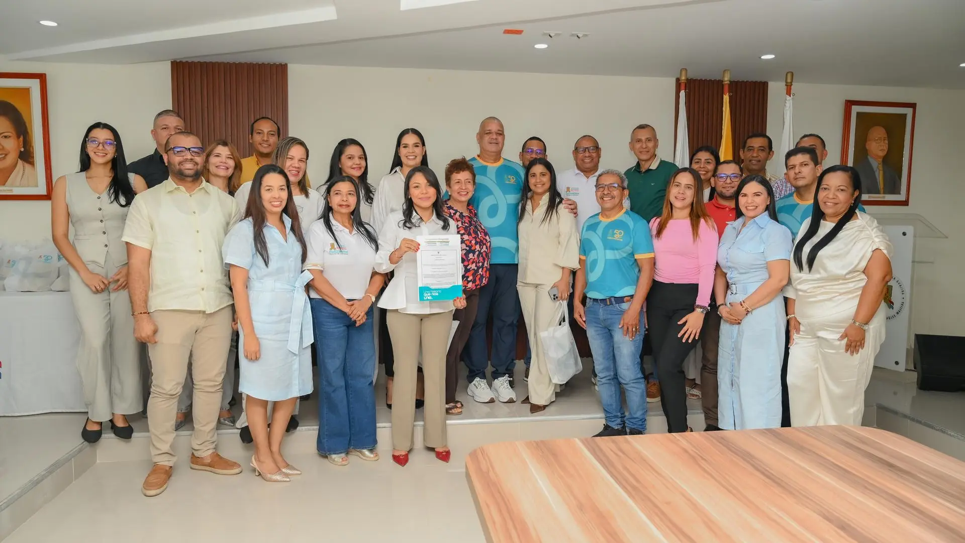 Fotografía grupal de funcionarios y colaboradores de la Universidad de La Guajira reunidos en una sala institucional. Varias personas posan frente a las banderas de la institución y de Colombia; en el centro, una integrante sostiene un documento, mientras otros participantes portan prendas conmemorativas de los 50 años de la Universidad de La Guajira