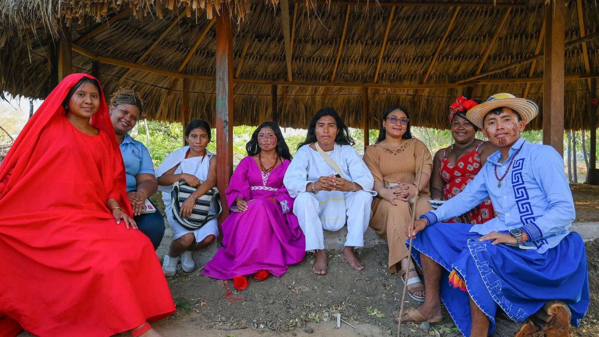 Fotografía de grupo de ocho personas posando juntas bajo un techo de paja. Visten ropas tradicionales indígenas en colores vibrantes: rojo, azul, marrón y tonos naturales, con accesorios, sombreros y adornos culturales tradicionales. Uno de los miembros sostiene un pequeño animal.