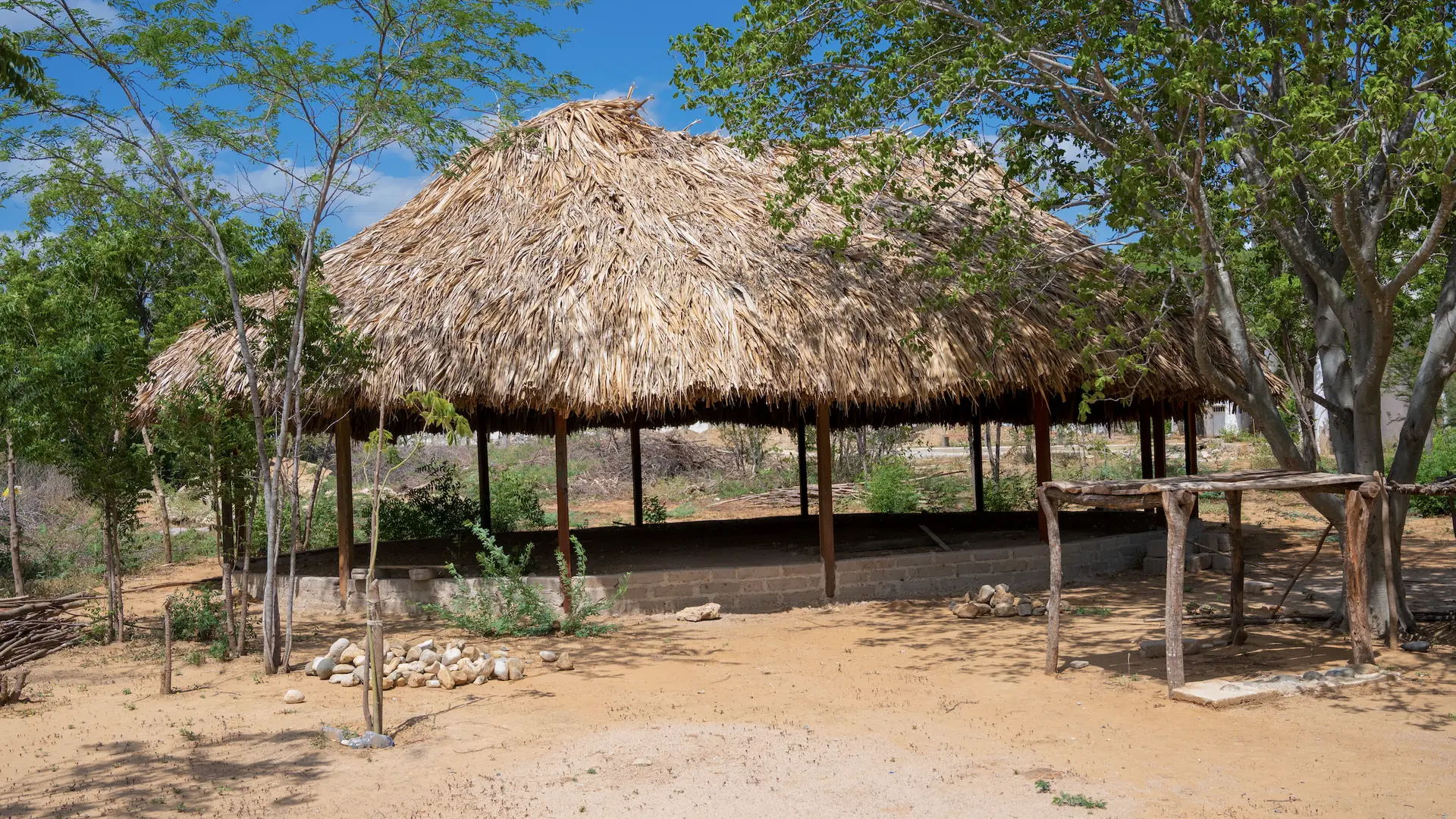 Grupo de cuatro personas sentadas bajo un techo de paja durante lo que parece ser una reunión comunitaria o sesión de capacitación. Las personas visten ropa tradicional e indígena con diseños y colores distintivos, algunos con accesorios y adornos tradicionales. Sostienen documentos y materiales educativos.