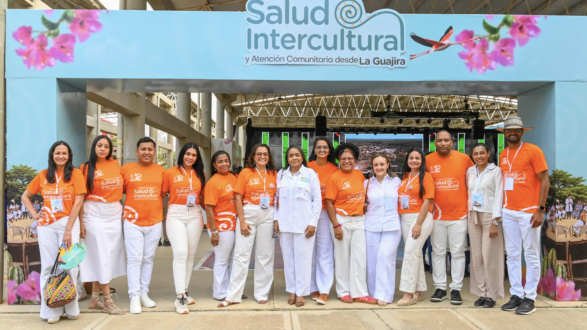 Un grupo de trece personas posa en fila sonriendo en un evento oficial. La mayoría viste camisetas naranjas con el logo "Salud Intercultural" y pantalones o faldas blancas. Al fondo destaca un gran letrero azul claro con la frase: "I Congreso Internacional de Salud Intercultural y Atención Comunitaria desde La Guajira", decorado con flores rosadas y la silueta de un flamenco.