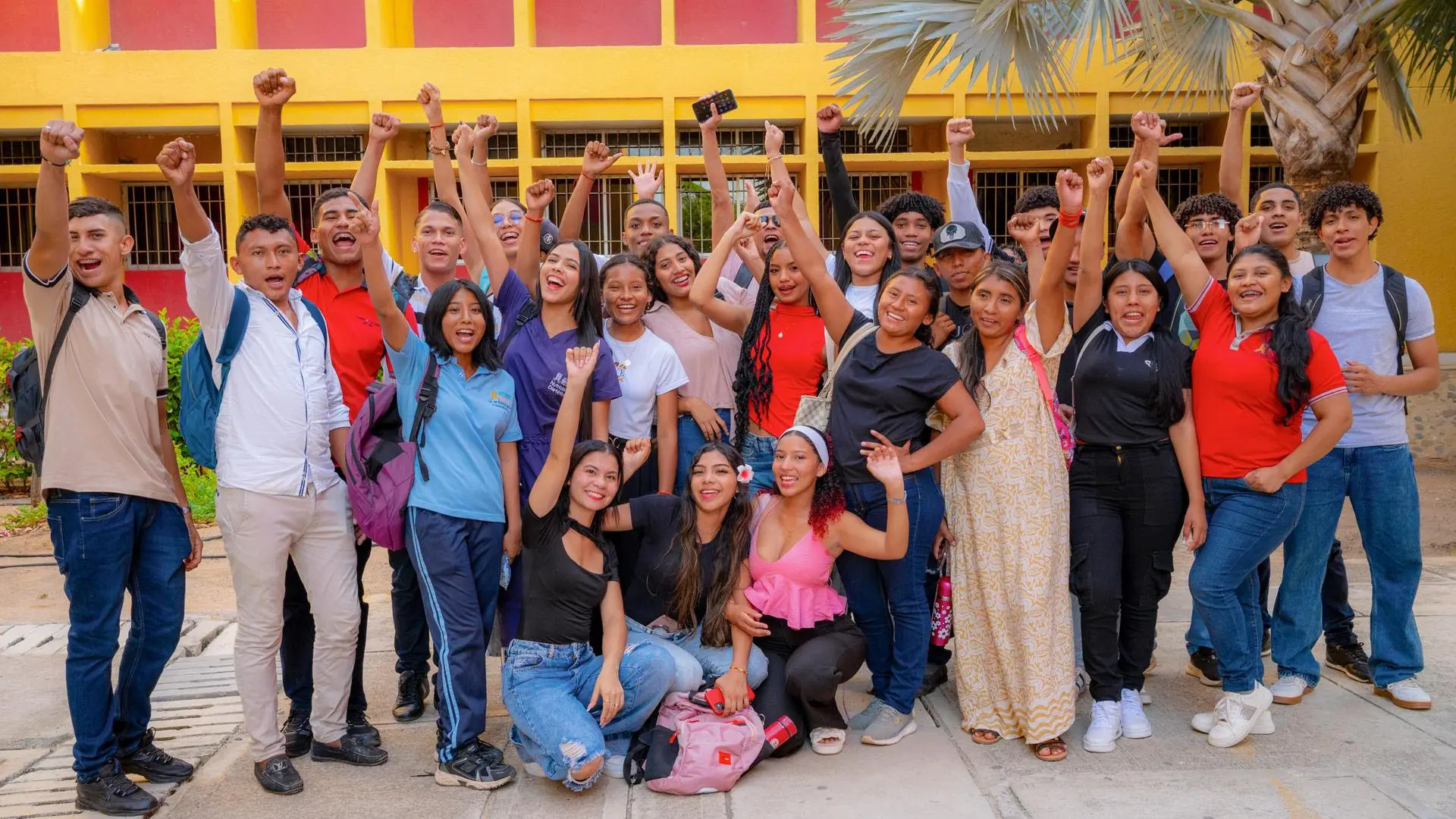 Grupo diverso de estudiantes posa frente a un edificio universitario de colores amarillo y rojo. Los jóvenes sonríen y levantan los brazos en señal de celebración, algunos llevan mochilas y prendas alusivas a la universidad.