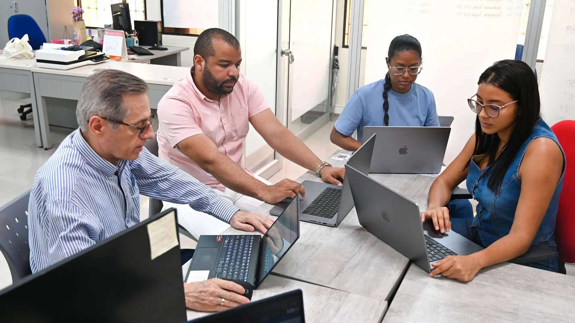 un grupo de cuatro personas trabajando en una oficina. Dos hombres y dos mujeres están sentados alrededor de una mesa, cada uno utilizando una computadora portátil. El hombre de mayor edad, a la izquierda, lleva una camisa de rayas y está usando un portátil ASUS. A su lado, otro hombre con camisa rosada señala la pantalla de su computadora. Las dos mujeres, una con camisa azul y la otra con un mono de mezclilla, también están concentradas en sus dispositivos Apple. El entorno es moderno y colaborativo, con escritorios organizados y equipos de trabajo.