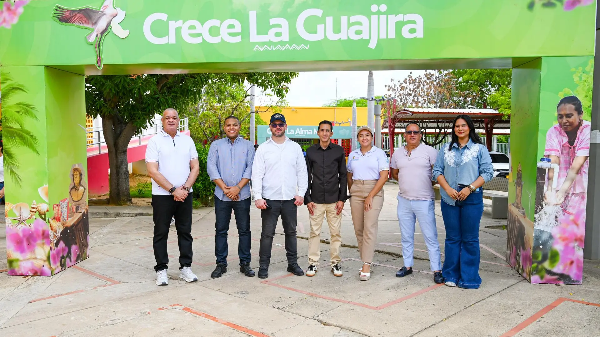 Grupo de siete personas posando de pie frente a un arco verde con la frase "Crece La Guajira".