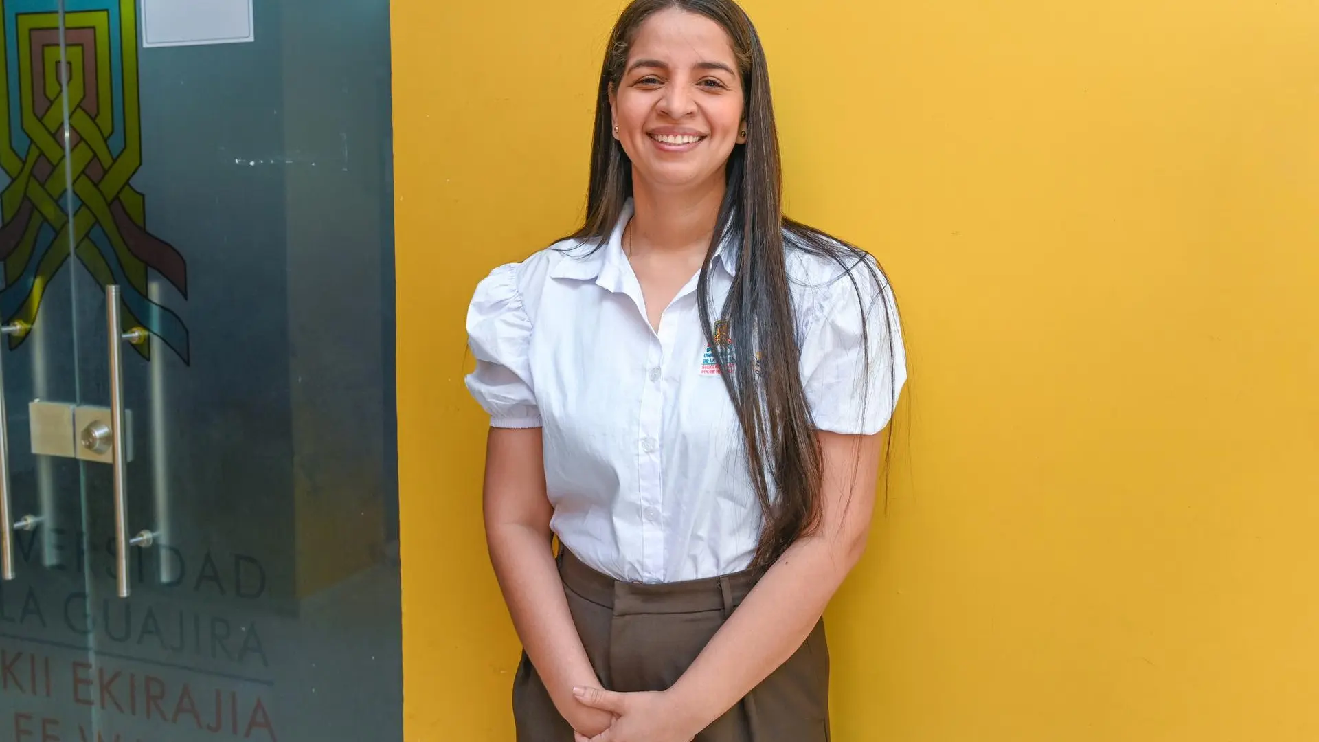 Mujer sonriente con camisa blanca institucional y pantalón café, de pie frente a una pared amarilla y una puerta de vidrio con el emblema de la Universidad de La Guajira.