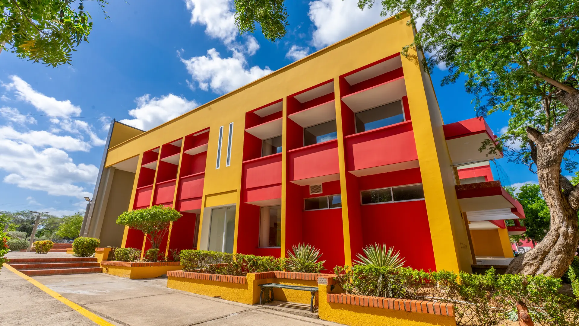 Fotografía de un edificio de la Universidad de La Guajira, con fachada amarilla y roja, rodeado de árboles y jardines, bajo un cielo azul con nubes blancas.