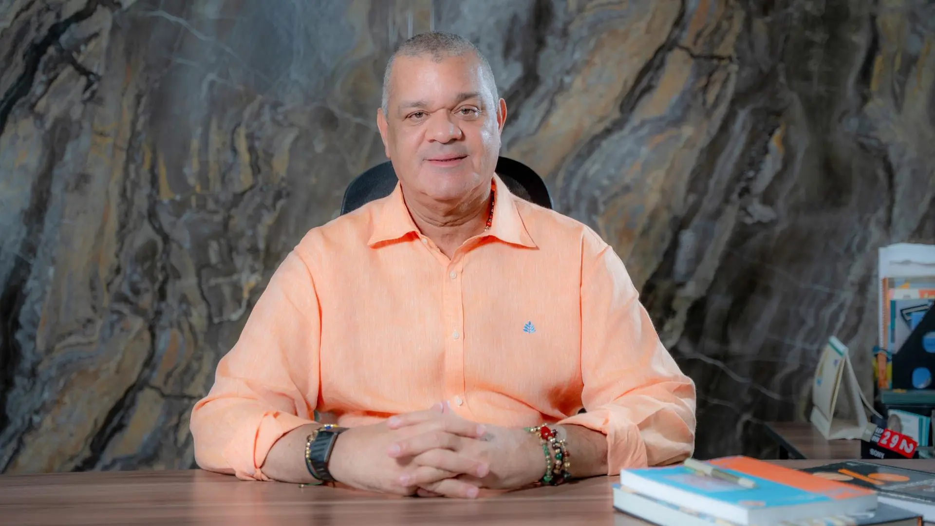 Fotografía de un directivo de la Universidad de La Guajira sentado detrás de un escritorio, con camisa naranja clara, en una oficina con pared de piedra y libros sobre la mesa.