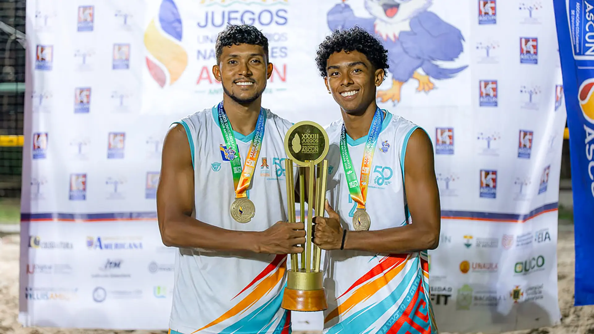 Heiber José Ureche Martínez y Yeiner José Vanegas Vergara, estudiantes de la Universidad de La Guajira, sonríen con sus medallas de oro y sostienen juntos el trofeo de campeones de voleibol playa. Visten el uniforme institucional y están frente al pendón de los XXXIII Juegos Nacionales Universitarios ASCUN.