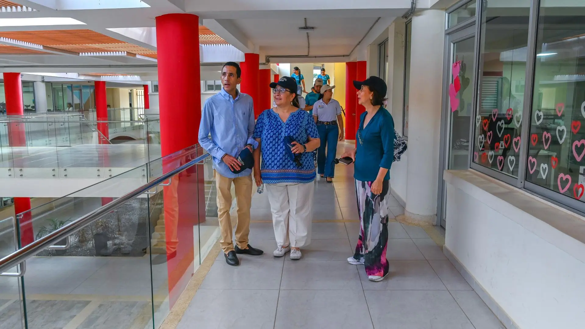 Tres personas conversan durante un recorrido por las instalaciones de la Universidad de La Guajira, en un pasillo con columnas rojas, barandas de vidrio y ventanales decorados con corazones de colores. Detrás de ellas, varias personas transitan por el corredor