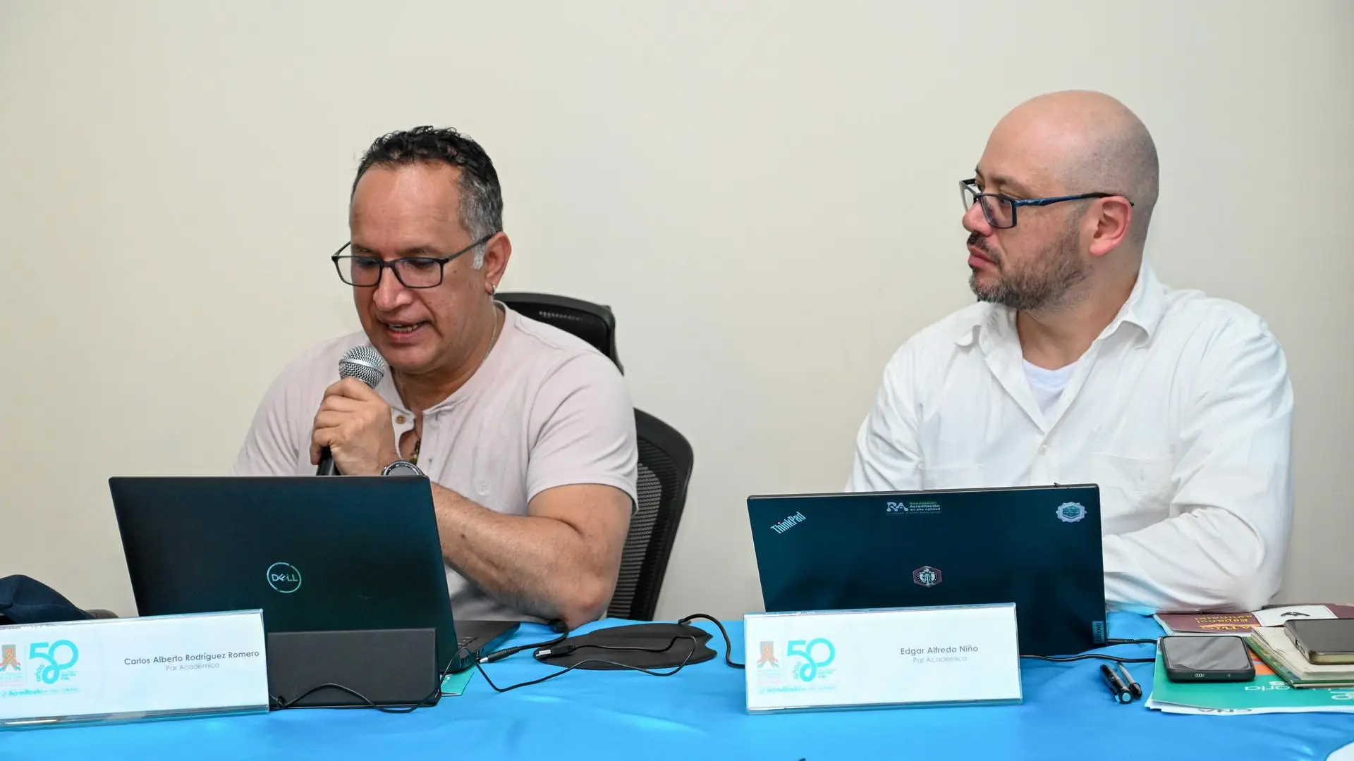Dos hombres sentados a una mesa con computadoras portátiles, uno de ellos hablando por un micrófono.