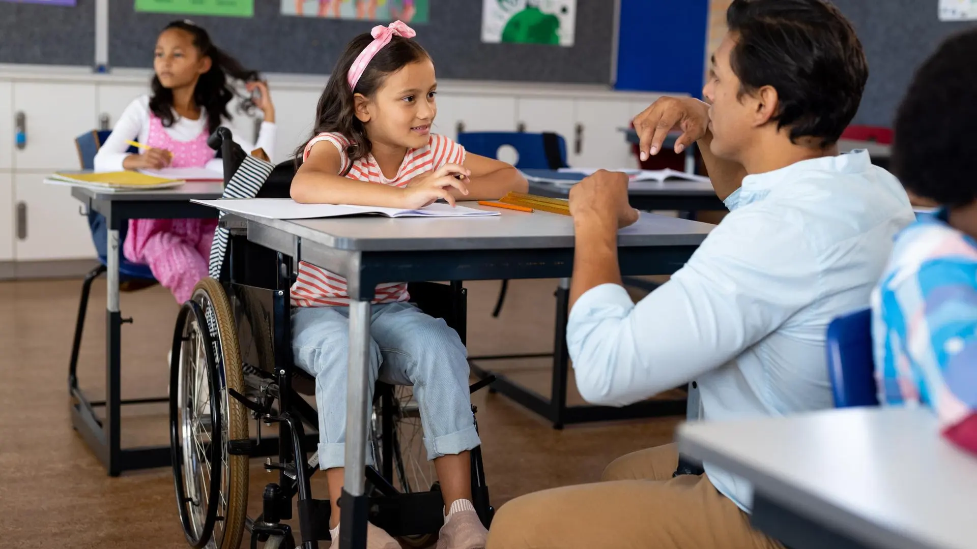 Docente conversa con una niña en silla de ruedas durante una actividad escolar en aula inclusiva.