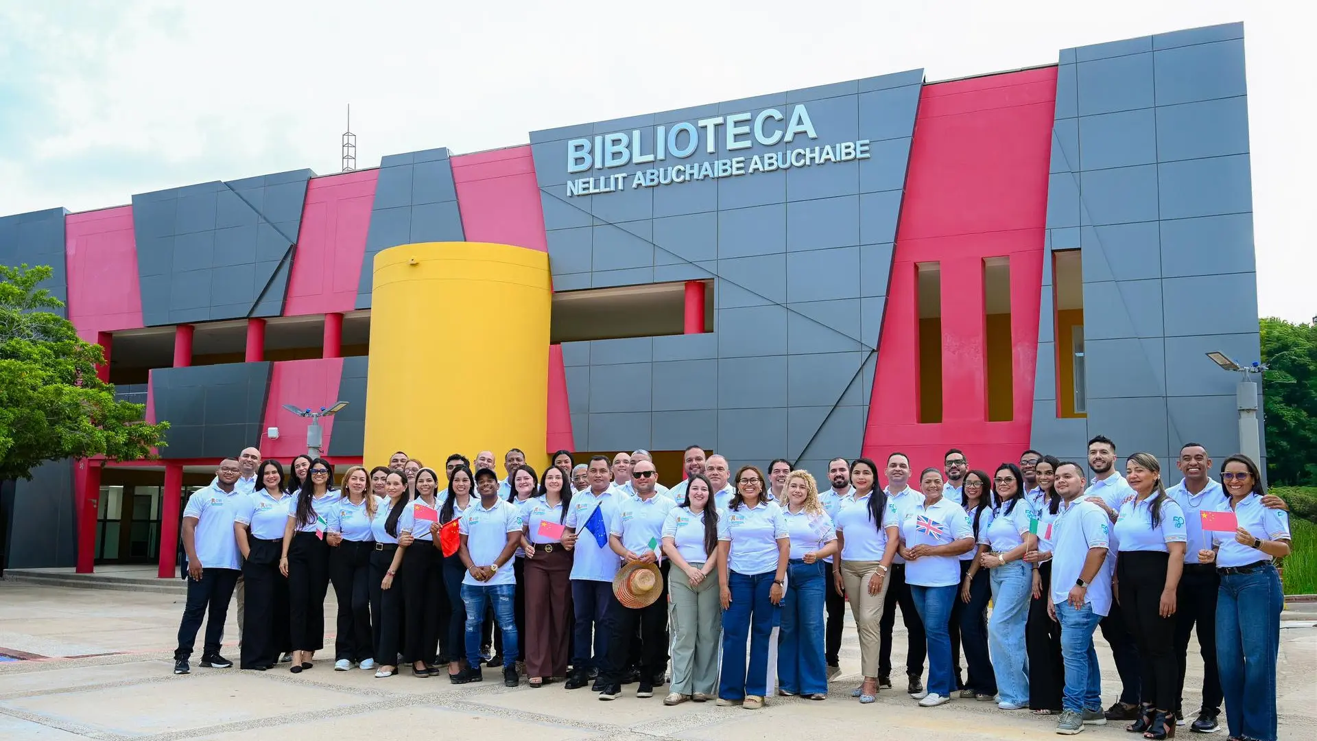 Grupo de docentes y participantes de Aula Global frente a la Biblioteca Nellit Abuchaibe Abuchaibe de la Universidad de La Guajira, sosteniendo banderas de diferentes países durante una jornada de intercambio académico e internacionalización.